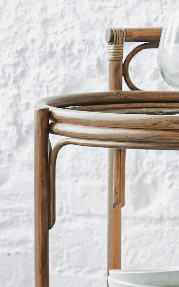Dette er et nærbillede af Romeo Rullebord i antique rattan. Detalje af det runde rattan-rullebordsstel mod en hvid murstensvæg.
