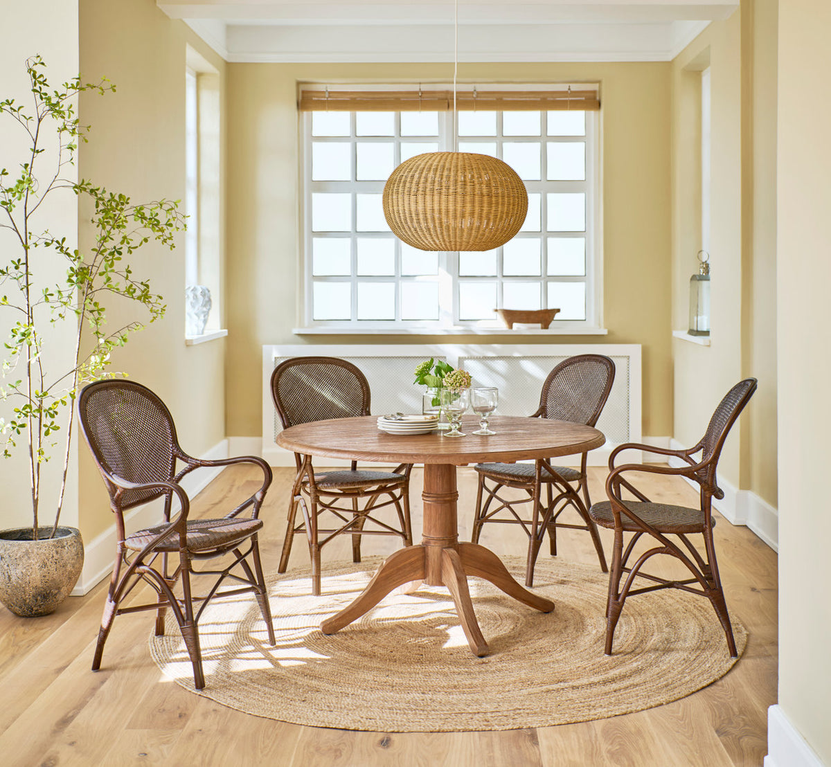 Dette er et livsstilsbillede af Rossini Spisestuelænestol i brown rattan. Rundt teakbord, rattanpendel og jutetæppe i en solrig spisestue.