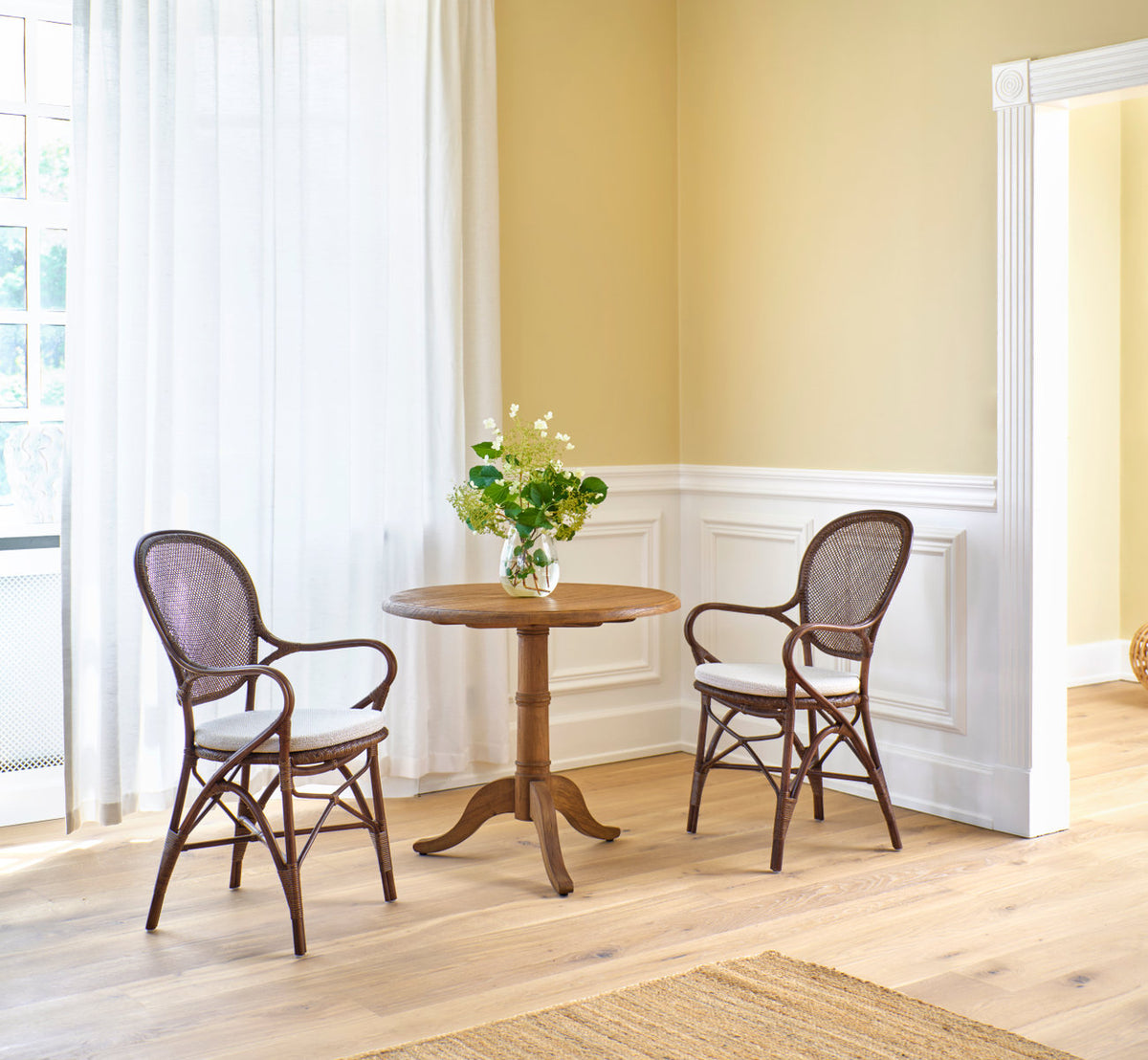 Dette er et livsstilsbillede af Rossini Spisestuelænestol i brown rattan. To ved et teak cafébord og en hvid panelvæg.