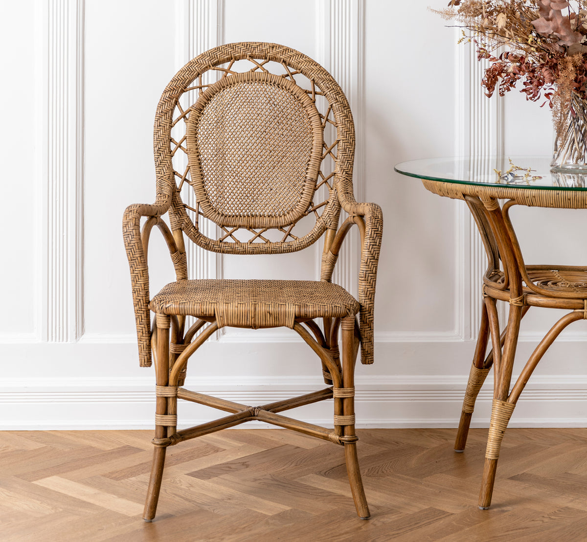Dette er et livsstilsbillede af Romantica Spisebordsstol i antique rattan. Placeret ved et glasbord i rattan med tørrede blomster i et panelrum.
