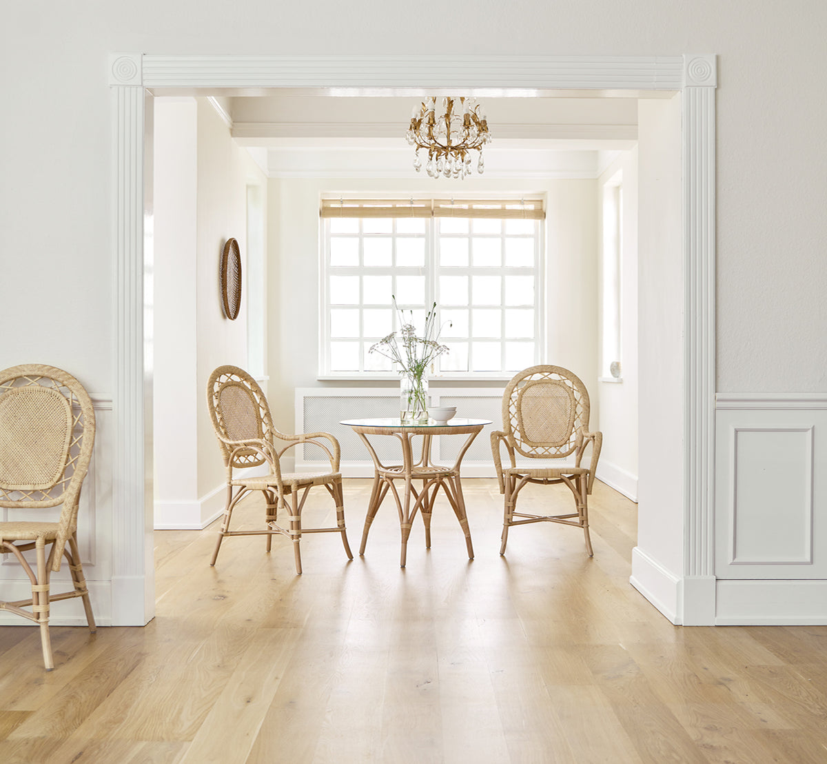Dette er et livsstilsbillede af Romantica Spisebordsstol i natural rattan. Omkring et rundt bord i et elegant rum med lysekrone og hvide paneler.