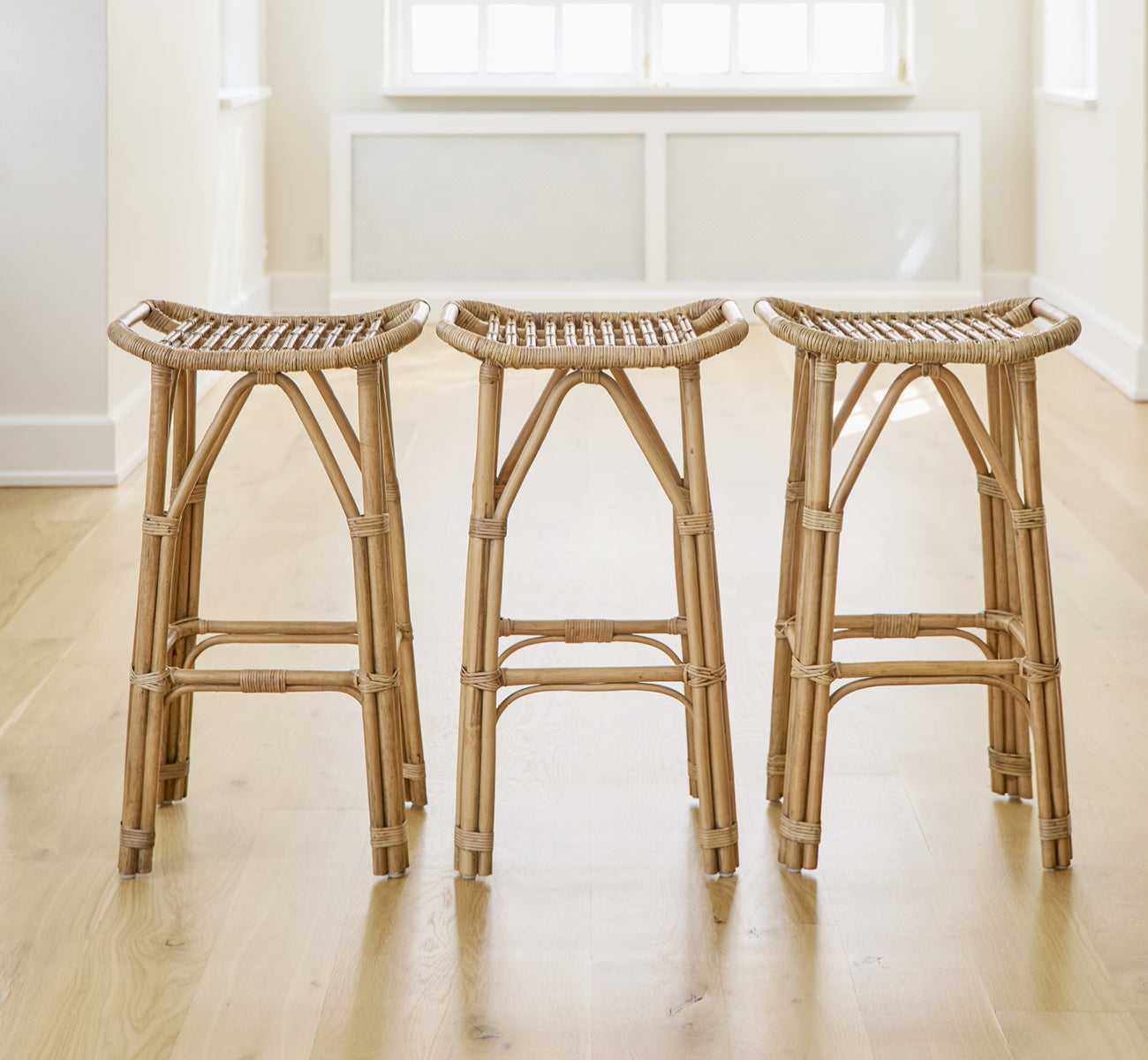 Dette er et livsstilsbillede af Salsa Barstol i antique rattan. Et par placeret mod en hvid væg på et sildebensparketgulv.