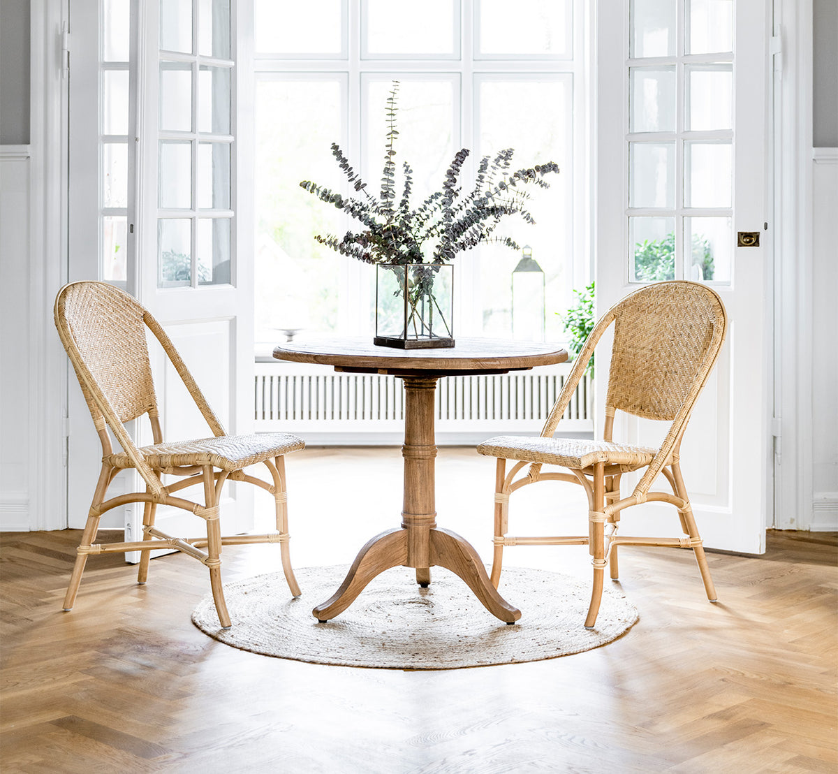 Dette er et livsstilsbillede af Alanis Spisebordsstol i natural rattan, i et lyst interiør med hvide panelvægge og sildebensgulv.