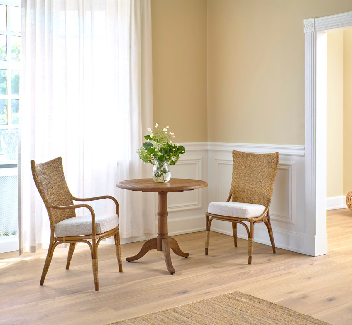Dette er et livsstilsbillede af Melody Spisestuelænestol i antique rattan. To ved et teak cafébord ved hvide paneler.