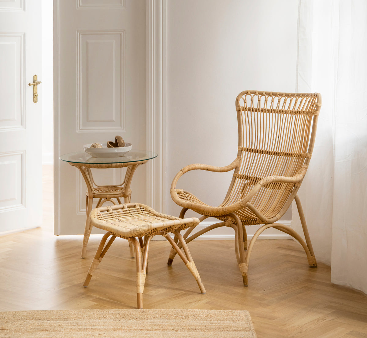 Dette er et livsstilsbillede af Monet Lounge Chair i natural rattan. Med fodskammel og sidebord nær en døråbning på sildebensgulv.