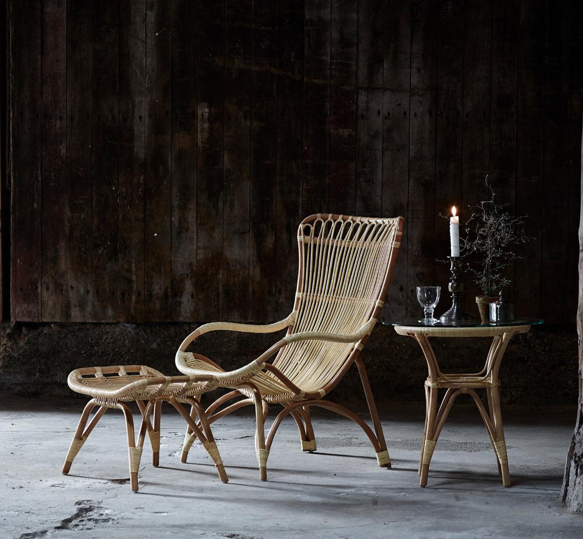 Dette er et livsstilsbillede af Monet Lænestol i natural rattan. Kombineret med en fodskammel og sidebord mod en mørk træbeklædt væg.