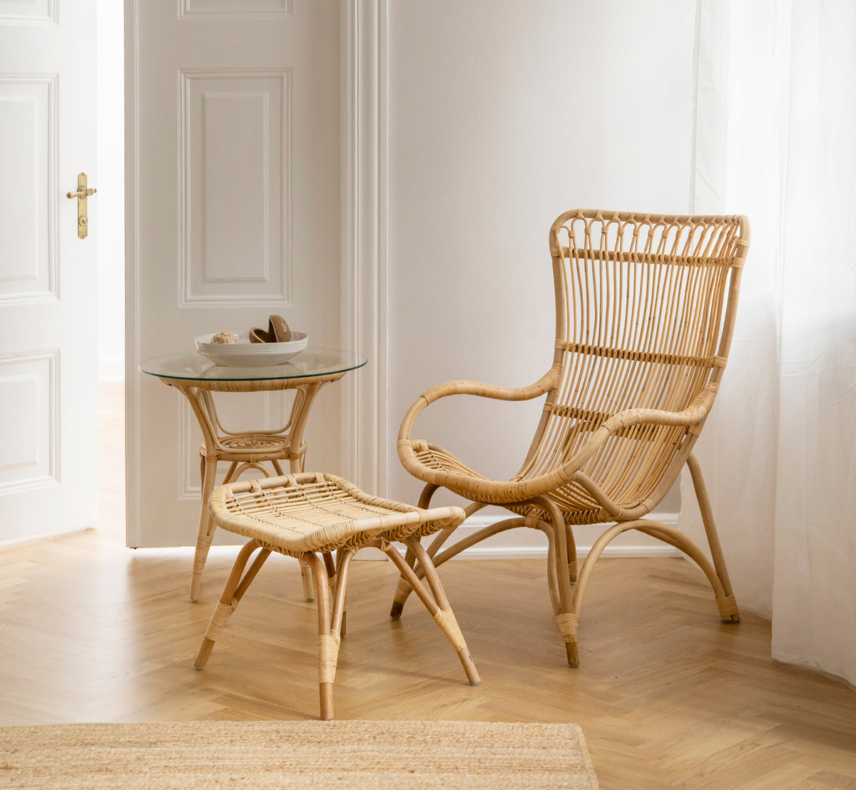 Dette er et livsstilsbillede af Monet Fodskammel i natural rattan. Kombineret med en højrygget rattanstol og glassidebord i en lys entré.