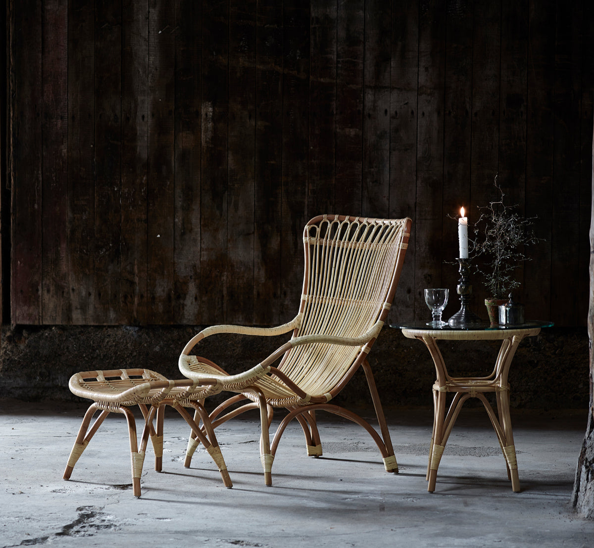Dette er et livsstilsbillede af Monet Footstool i natural rattan. Med lænestol i en mørk rustik ladescene med glasvarer.