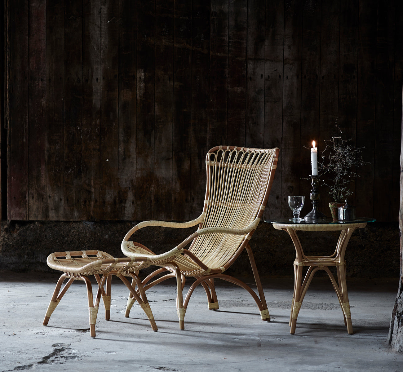 Dette er et livsstilsbillede af Monet Footstool i natural rattan. Med lænestol i en mørk rustik ladescene med glasvarer.