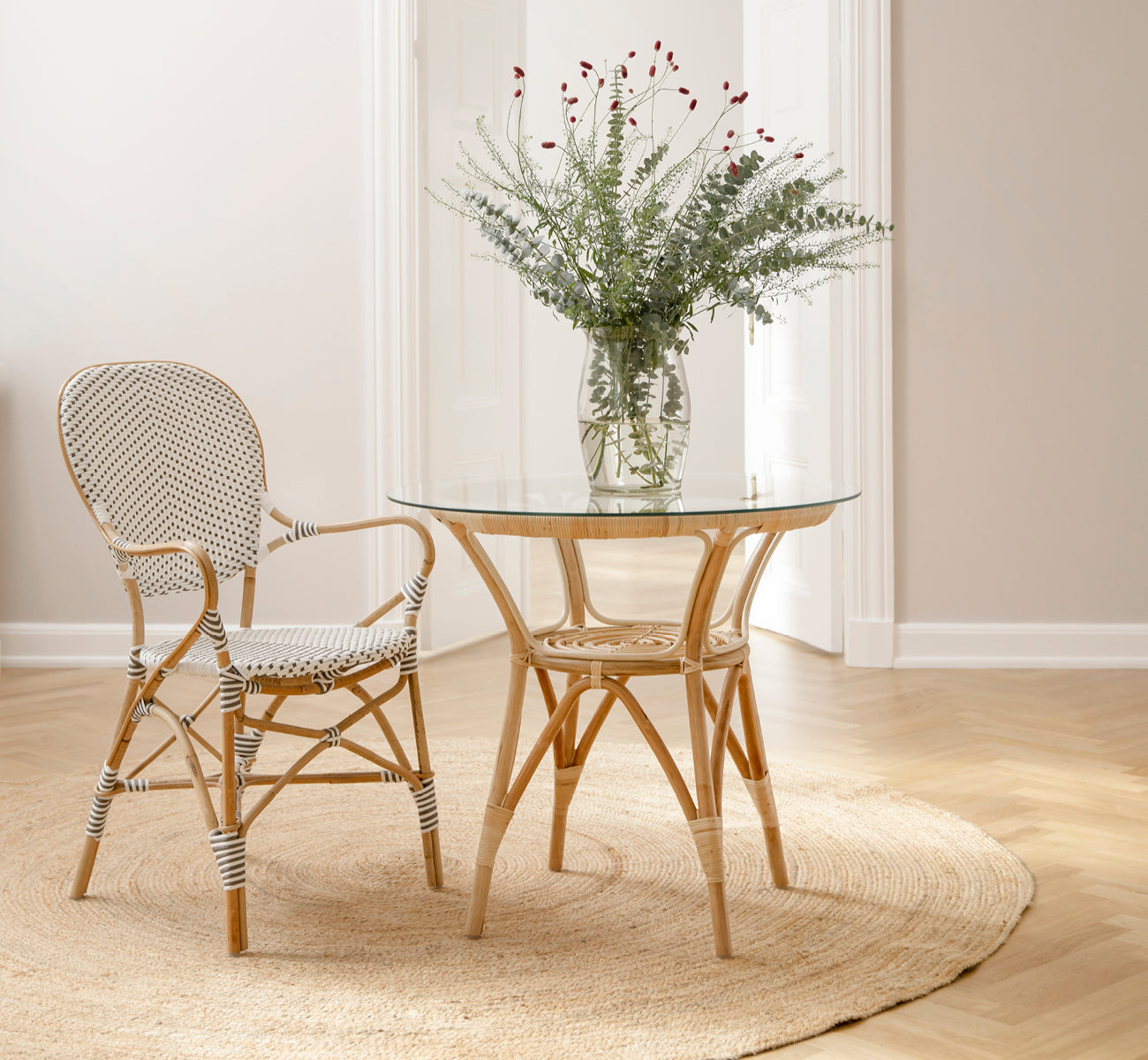 Dette er et livsstilsbillede af Tony Café Table i natural rattan. Kombineret med to stole på et rundt jutetæppe i en lys døråbning.