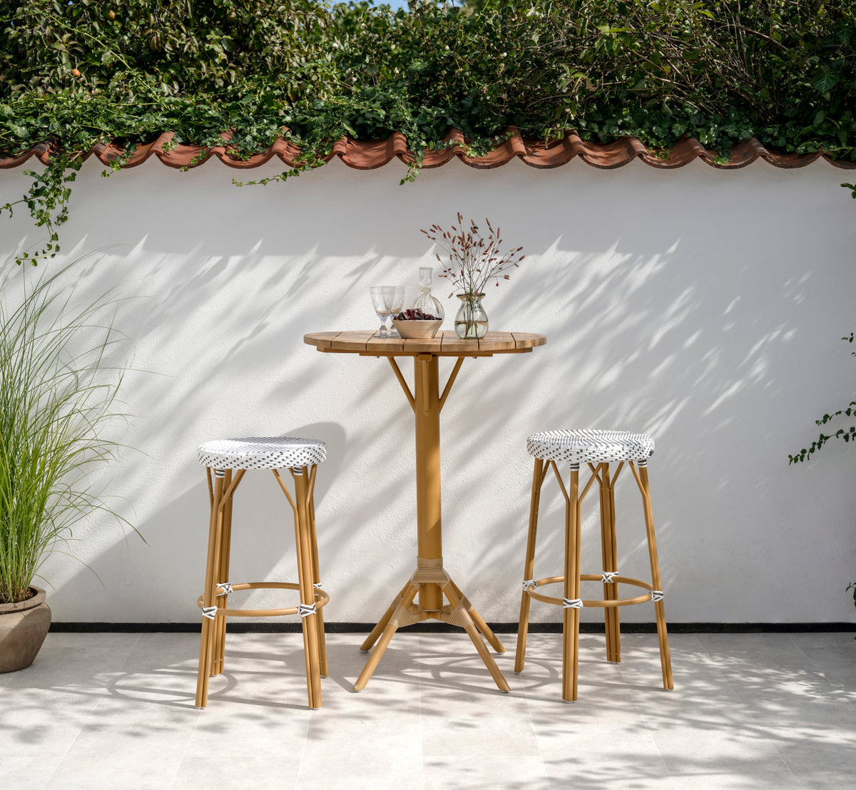 Dette er et livsstilsbillede af Simone Udendørs barstol i almond aluminium frame white w cappuccino dot artfibre. Ved et teak barbord på en terrasse.