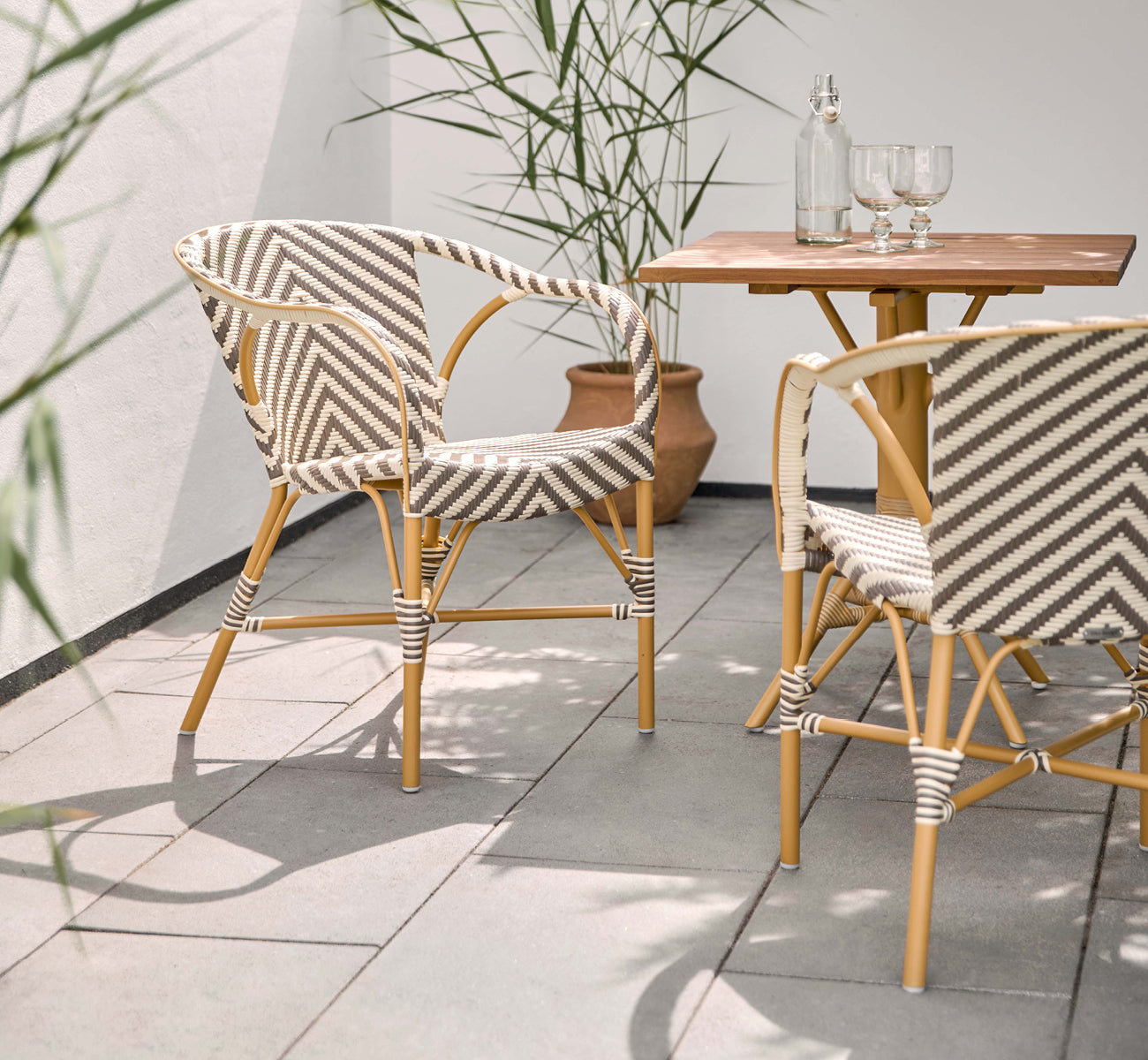 Dette er et livsstilsbillede af Madeleine Udendørs spisebordsstol i almond aluminum frame chevron cappuccino ivory ArtFibre. Solrig terrasse med teakbord.