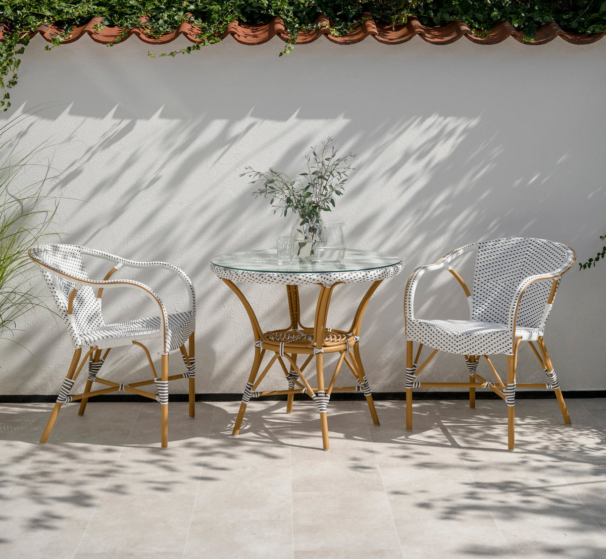 Dette er et livsstilsbillede af Madeleine Udendørs spisebordsstol i almond aluminium frame white w cappuccino dot artfibre. Ved et glasbord på en terrasse.