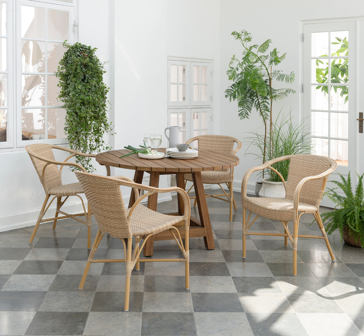 Dette er et livsstilsbillede af Madeleine Udendørs spisebordsstol i almond aluminium frame natural ArtFibre. Lys orangeri med planter og flisegulv.