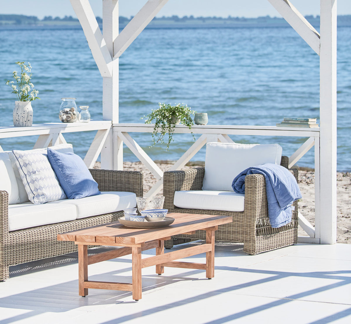 Dette er et livsstilsbillede af Carrie Lænestol i antique Grey ArtFibre Aluminium, på en soldrenkt kystterrasse med havudsigt og hvid pergola.