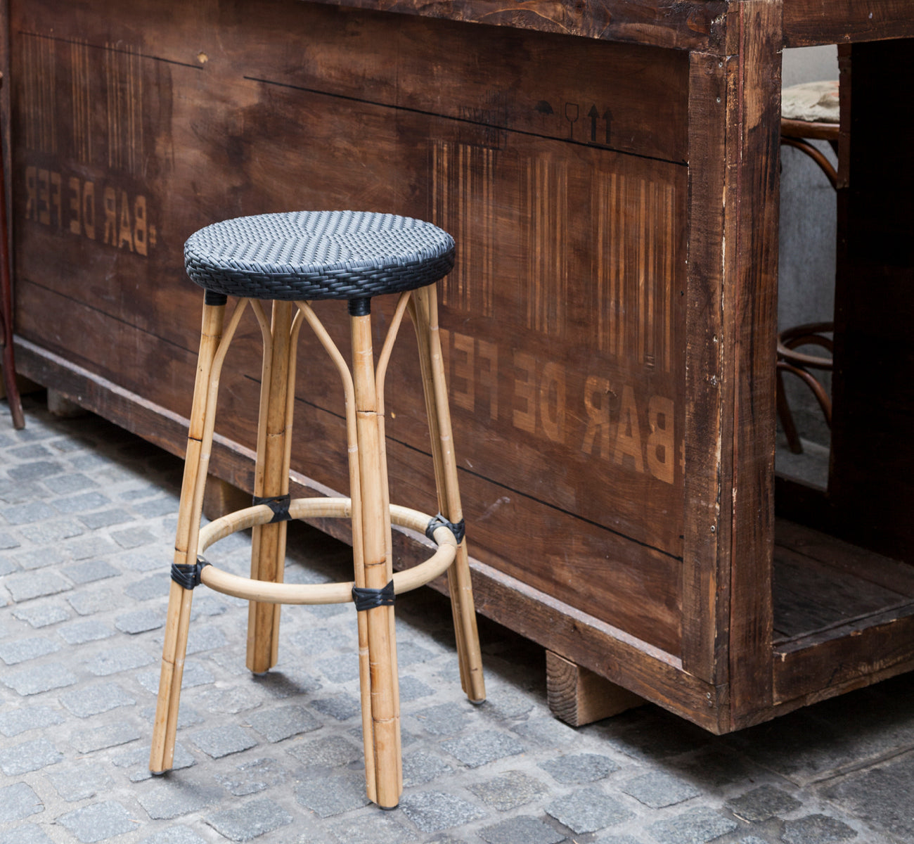 Dette er et livsstilsbillede af Simone Barstol i black ArtFibre Skin on Rattan frame. Placeret ved en rustik træbar på brosten.