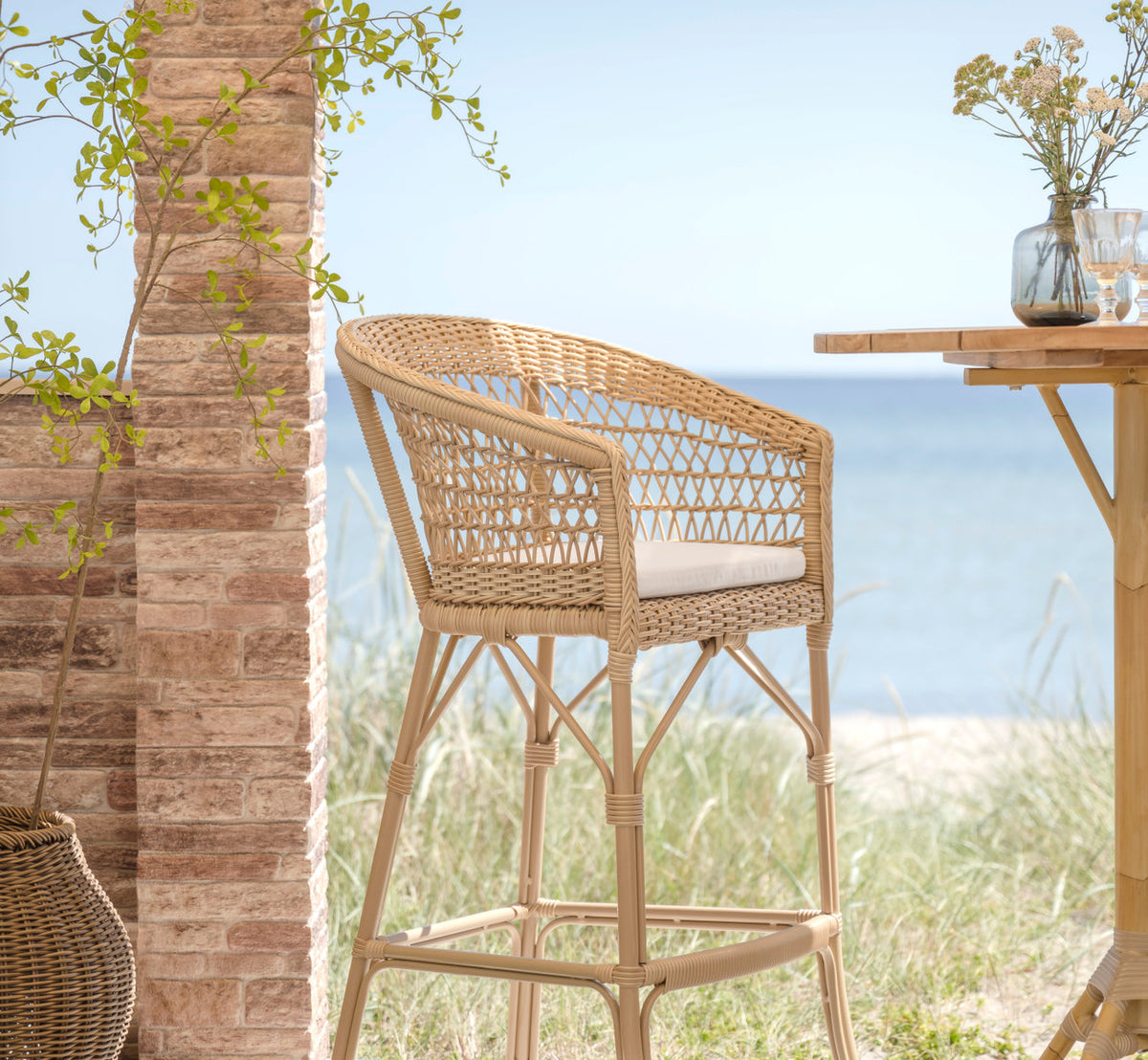 Dette er et livsstilsbillede af Emma Udendørs barstol i natural artfibre aluminium. Ved en murstenssøjle med klittergræs og havudsigt.