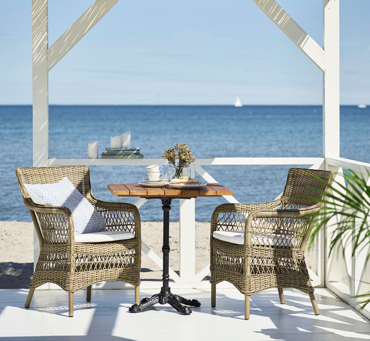 Dette er et livsstilsbillede af Marie Exterior Armchair i antique grey artfibre aluminium. Ved et teakbord i en strandpavillon.