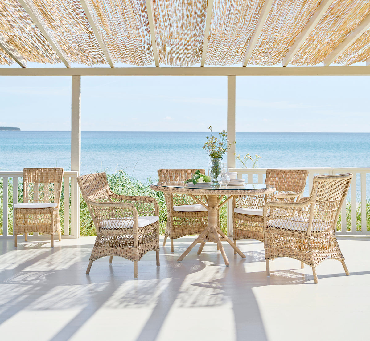 Dette er et livsstilsbillede af Marie Exterior Armchair i natural artfibre aluminium. På en overdækket veranda med havudsigt.