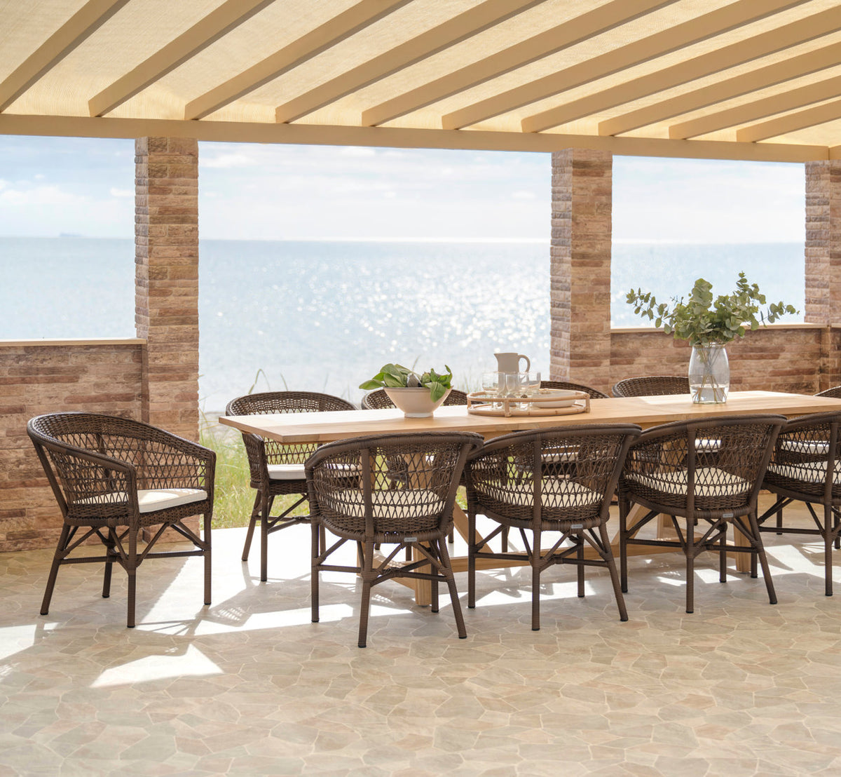 Dette er et livsstilsbillede af Emma Udendørs stol i brown artfibre aluminum. Langt spisebord dækket på en terrasse ved havet.