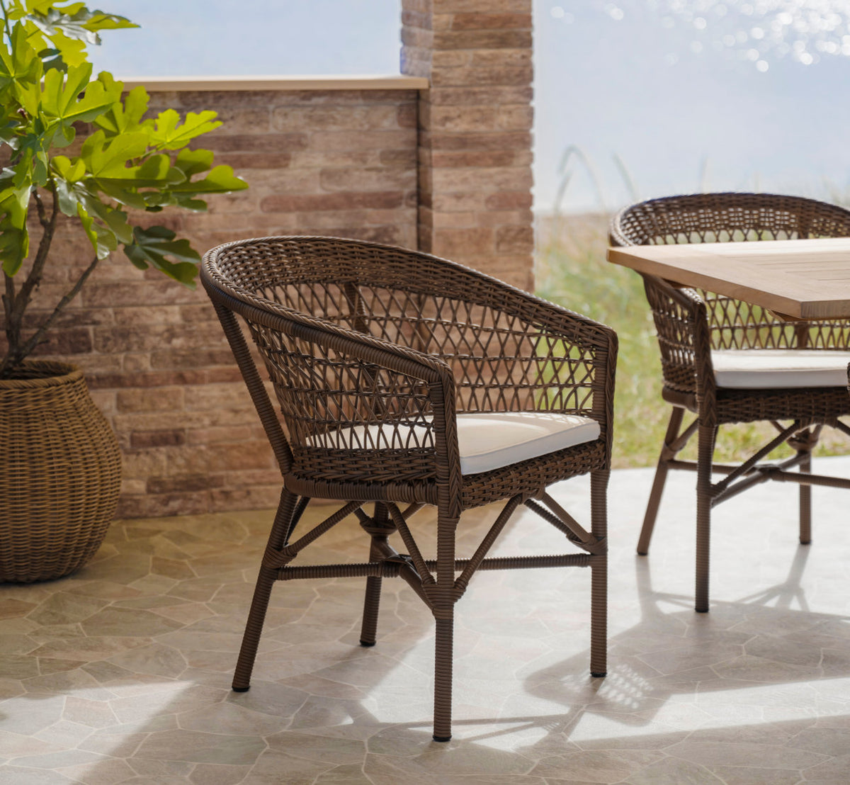 Dette er et livsstilsbillede af Emma Udendørs stol i brown artfibre aluminum. Ved en solbeskinnet murstensvæg på en terrasse.