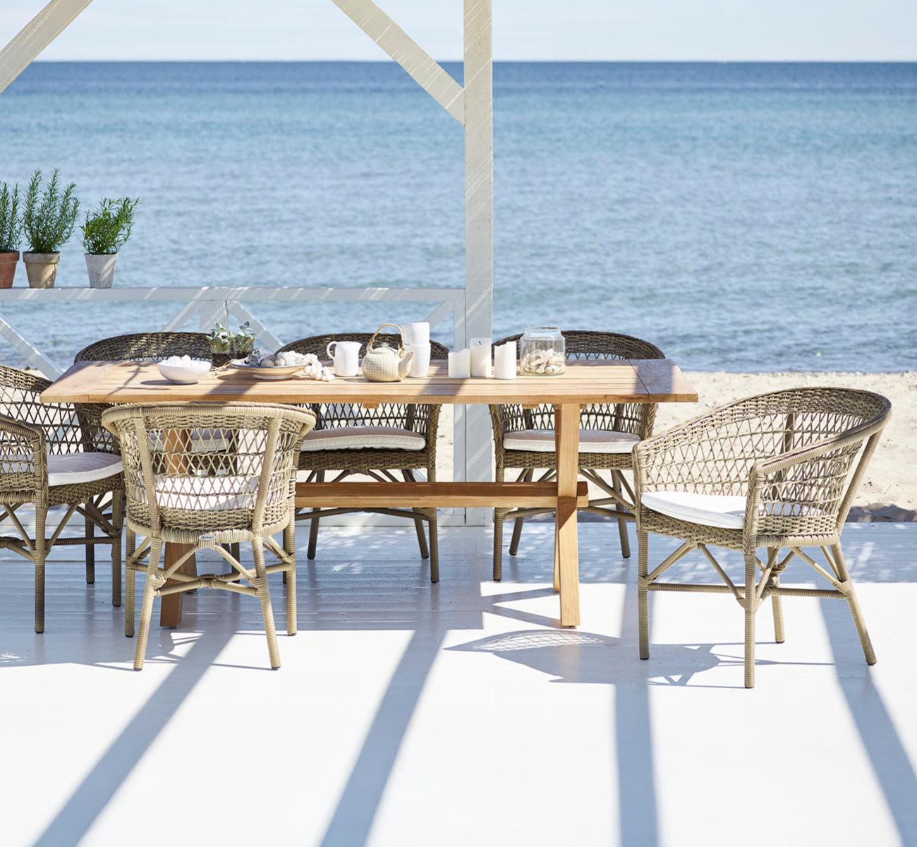 Dette er et livsstilsbillede af Emma Udendørs Spisebordsstol i antique Grey ArtFibre Aluminium, på en lys kystterrasse med sivtag og havudsigt.