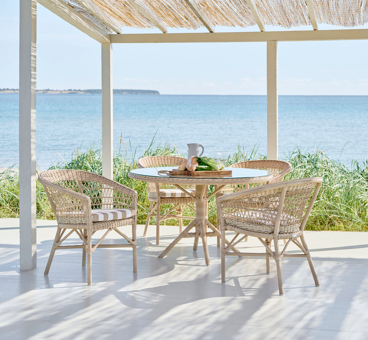 Dette er et livsstilsbillede af Emma Udendørs Spisebordsstol i natural ArtFibre Aluminium, på en lys kystterrasse med sivtag og havudsigt.