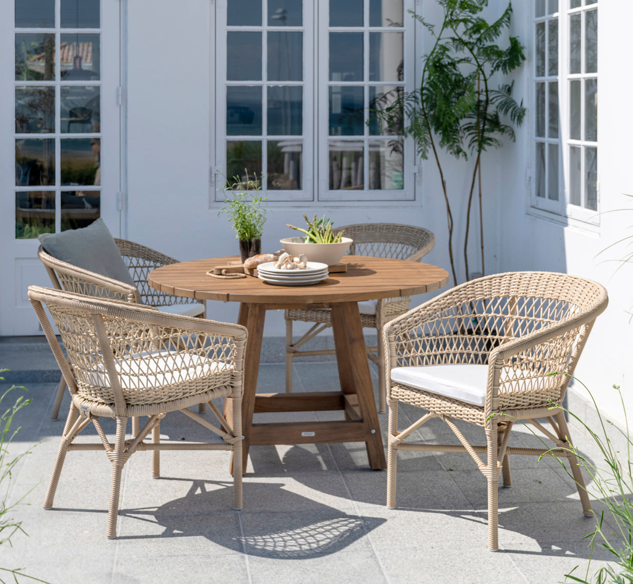 Dette er et livsstilsbillede af Emma Udendørs Spisebordsstol i natural ArtFibre Aluminium, på en lys kystterrasse med sivtag og havudsigt.
