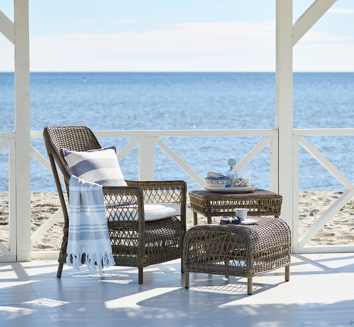 Dette er et livsstilsbillede af Dawn Lænestol i antique Grey ArtFibre Aluminium, på en kystterrasse med havudsigt, strandgræs og hvide søjler.