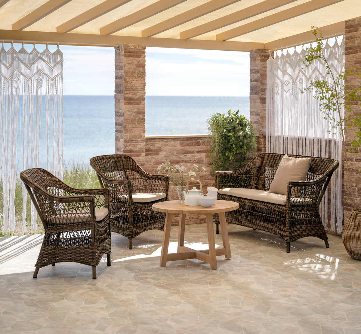 Dette er et livsstilsbillede af Charlot 2-personers udendørs sofa i brown artfibre aluminum. Med teak sofabord og makramégardiner ved havet.
