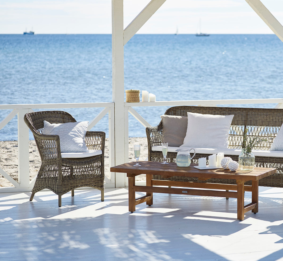 Dette er et livsstilsbillede af Charlot 3-personers sofa i antique Grey ArtFibre Aluminium, på en kystterrasse med sivtag og havudsigt.