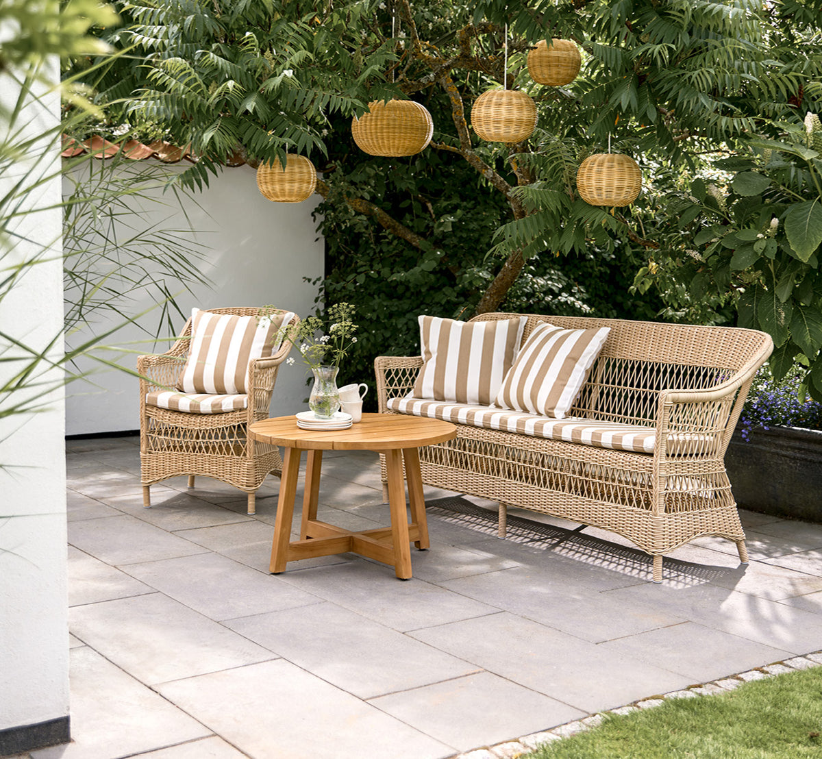 Dette er et livsstilsbillede af Charlot 3-personers sofa i natural ArtFibre Aluminium, på en kystterrasse med sivtag og havudsigt.
