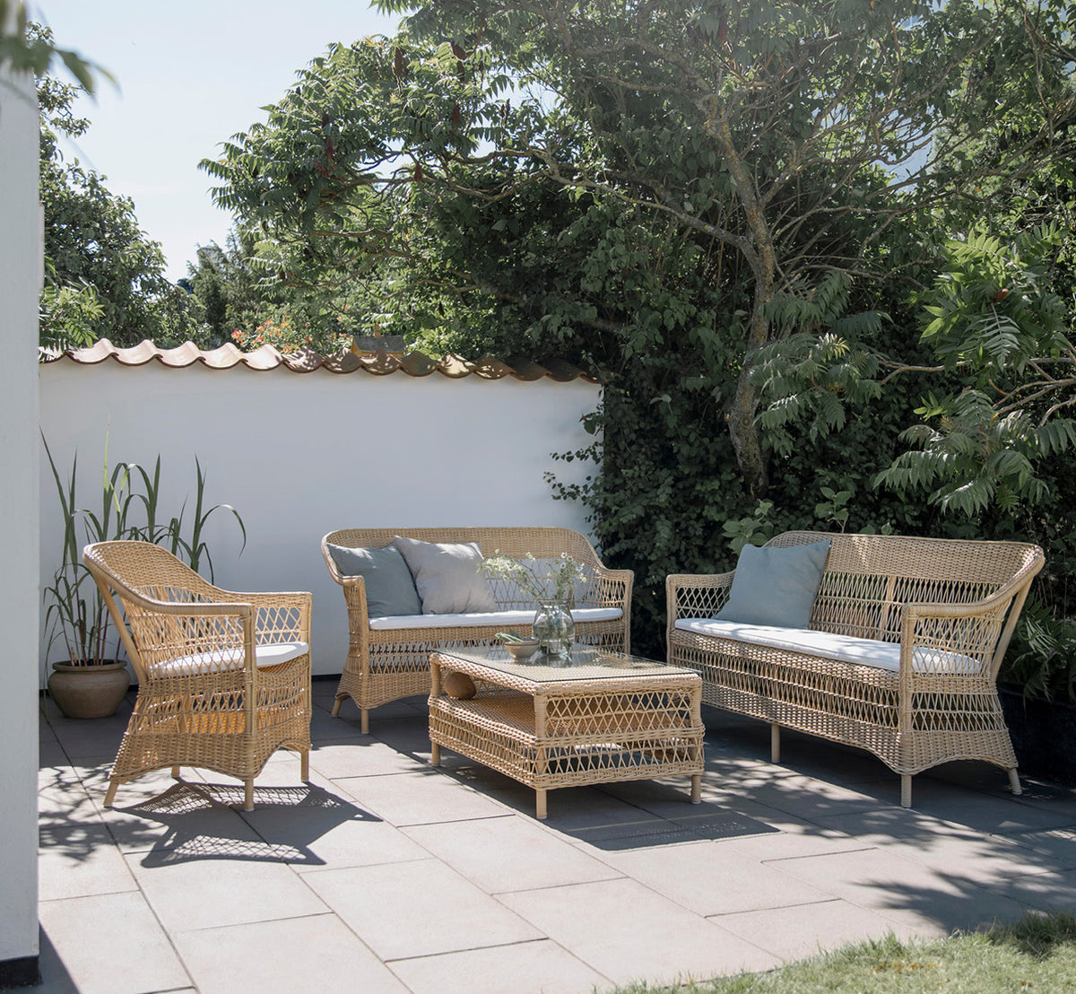 Dette er et livsstilsbillede af Charlot 3-personers sofa i natural ArtFibre Aluminium, på en kystterrasse med sivtag og havudsigt.
