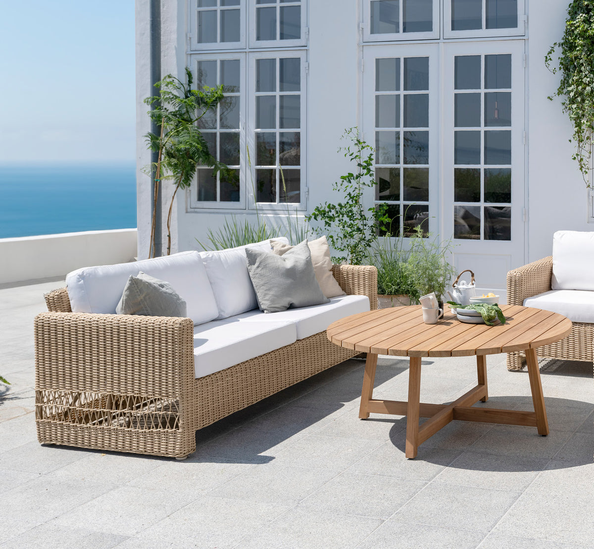 Dette er et livsstilsbillede af Carrie Sofa i natural ArtFibre Aluminium, på en kystterrasse med hvidt trædæk, havudsigt og pergola.
