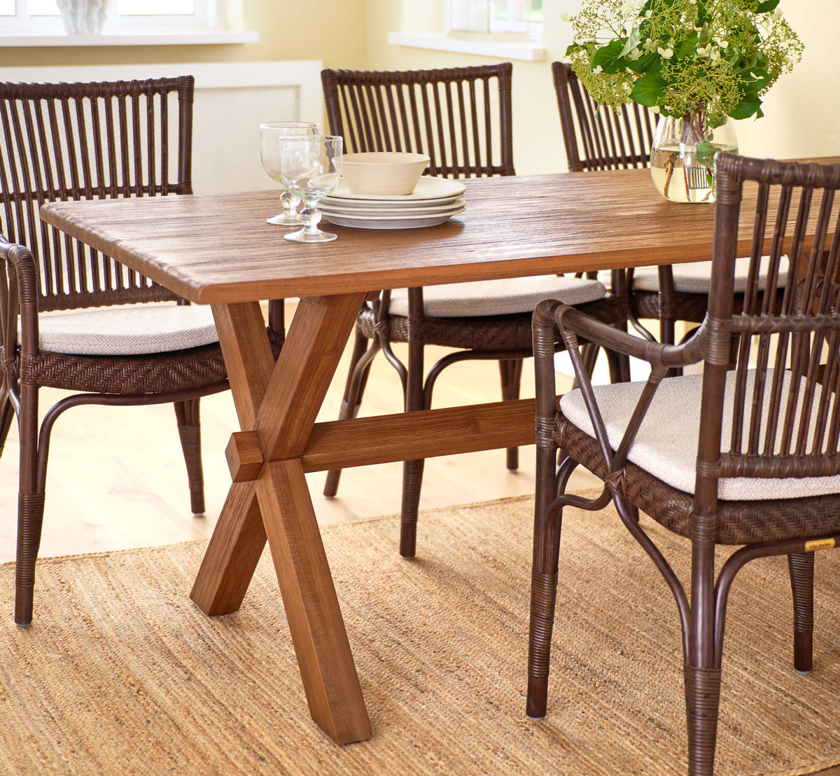 Dette er et livsstilsbillede af Hugo Teak spisebord 180x90 cm i teak indoor. Med rattan spisebordsstole på et jutetæppe.