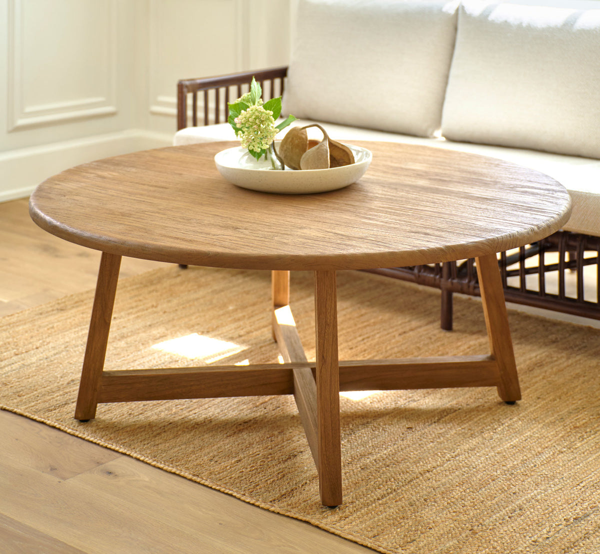 Dette er et livsstilsbillede af Aksel Sofabord Ø120 cm i teak indoor. Med en skål blomster foran en rattansofa.