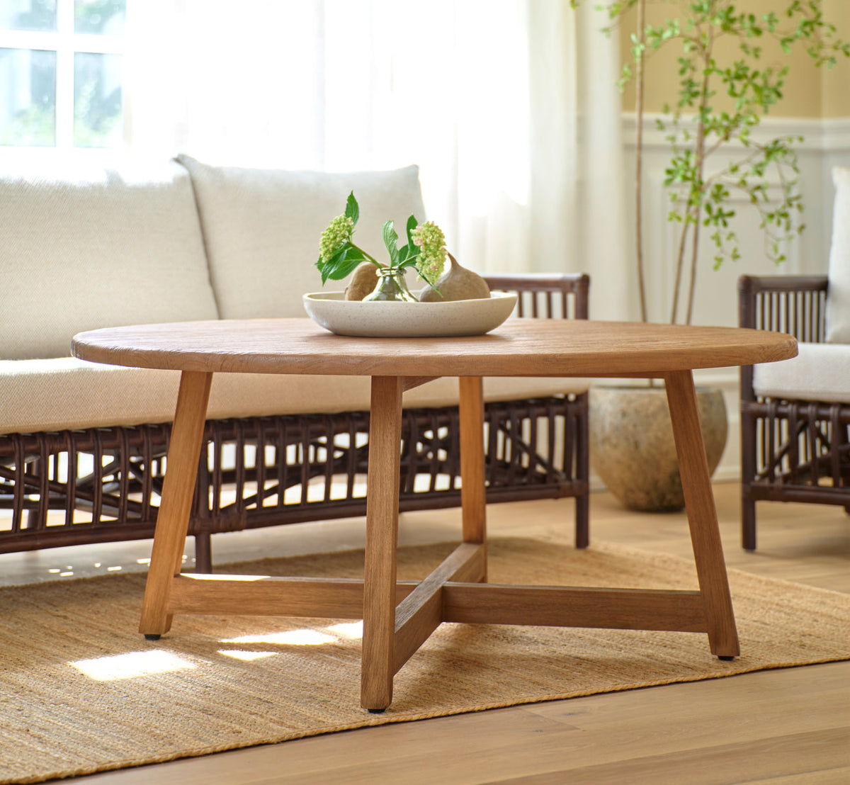 Dette er et livsstilsbillede af Aksel Sofabord Ø120 cm i teak indoor. Foran en rattansofa i en solrig stue.