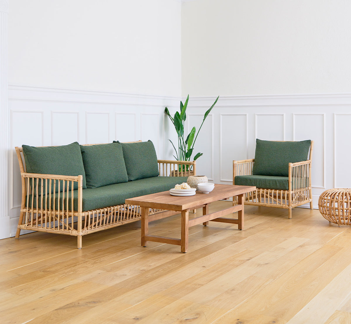 Dette er et livsstilsbillede af Alfred Sofabord i teak indoor, i en lys stue med hvide panelvægge og en sofa med grønne puder.