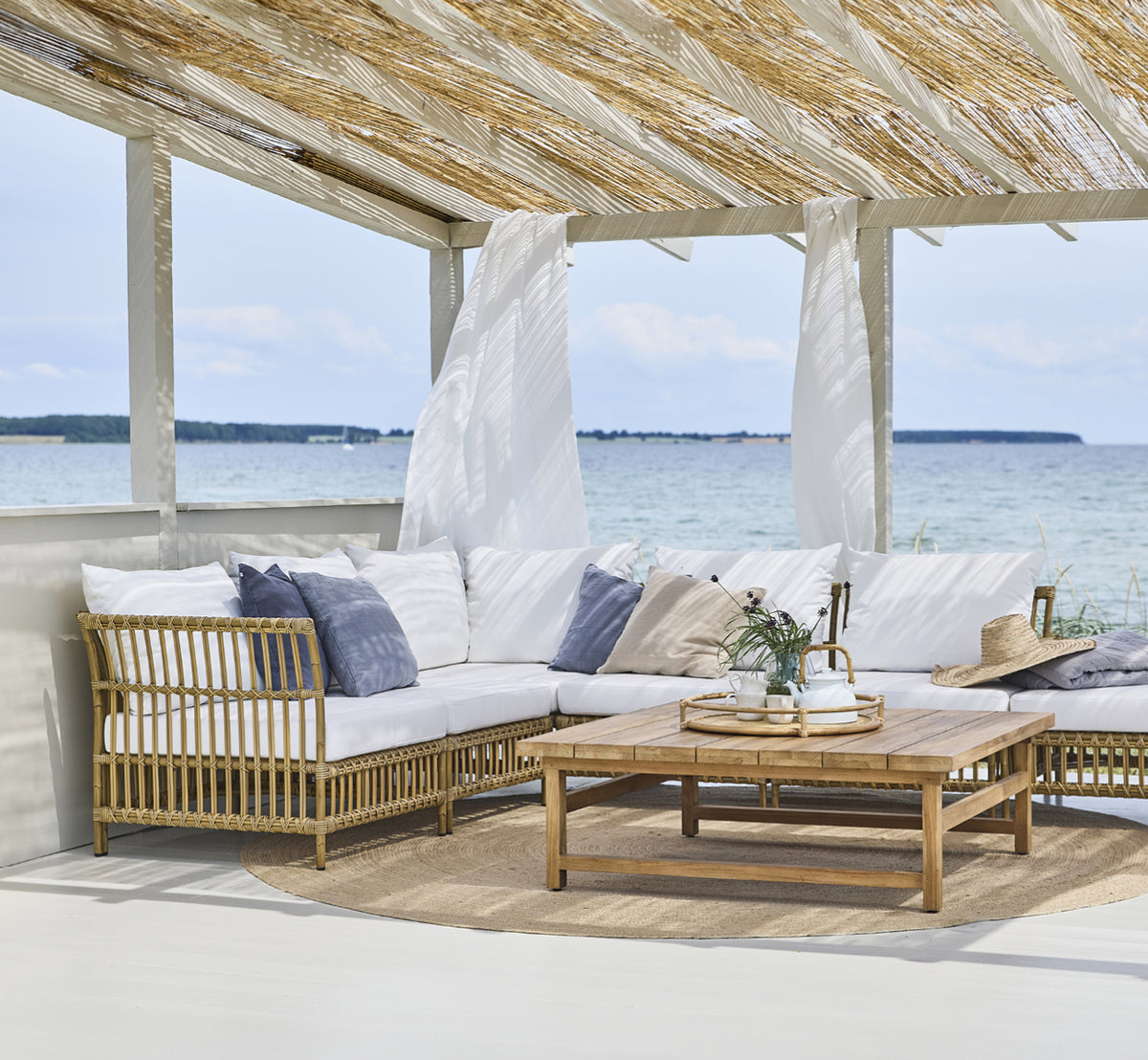 Dette er et livsstilsbillede af Julian Teak Lounge Table 120x120 cm i teak outdoor. Under en strandpergola med loungesæder og havudsigt.