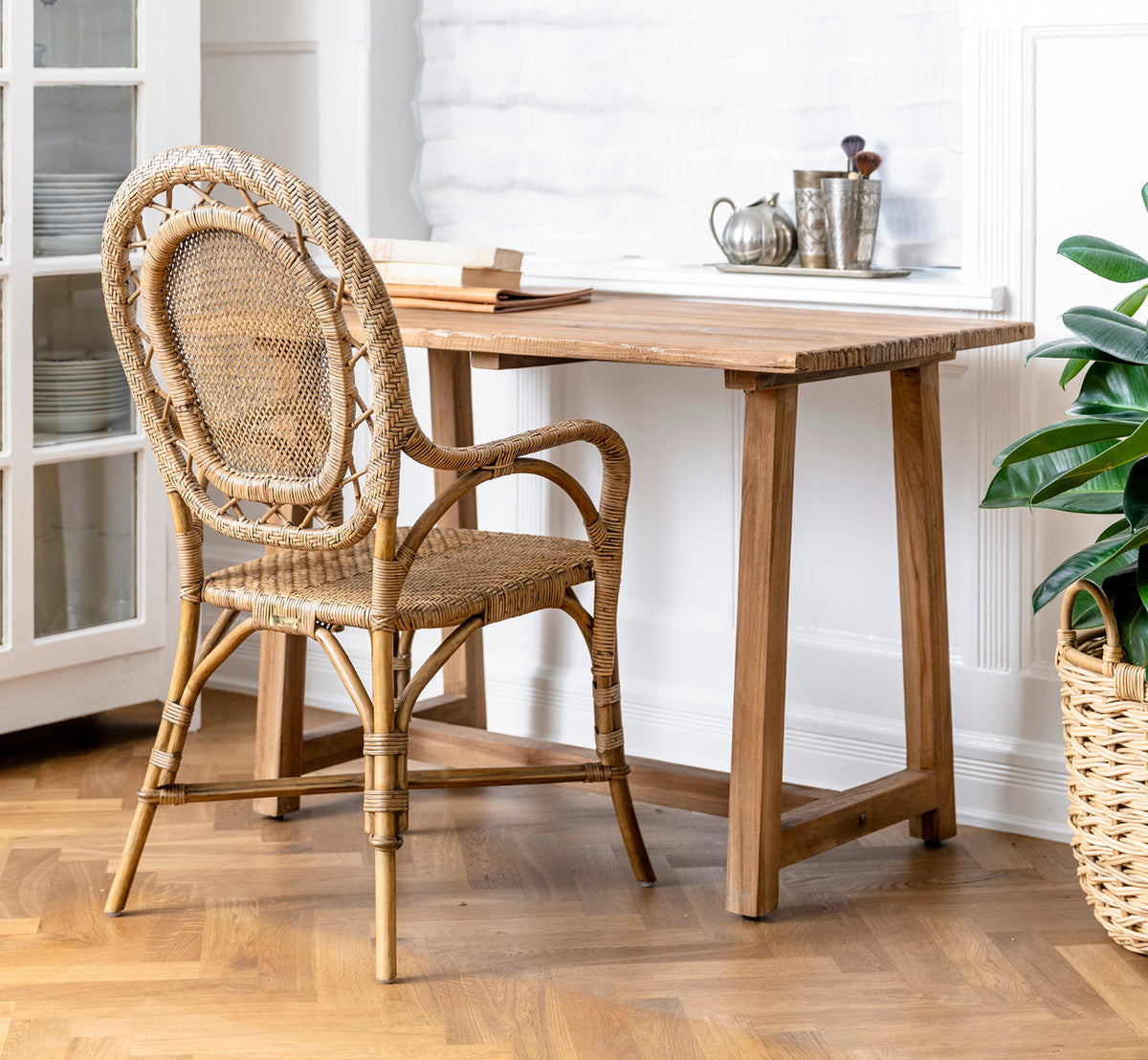 Dette er et livsstilsbillede af Lucas Teak Skrivebord i teak indoor. Kombineret med en rattanstol og grøn plante i et lyst interiør med sildebensgulv.