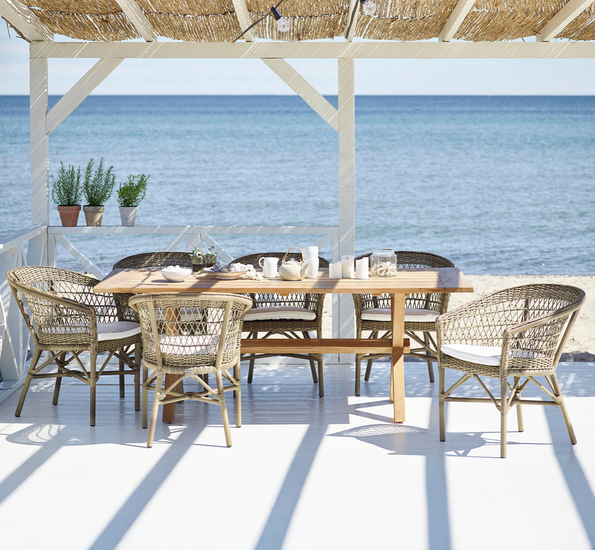 Dette er et livsstilsbillede af Colonial Bord 200 i outdoor Teak, på en strandterrasse med hvidt trædæk og havudsigt.