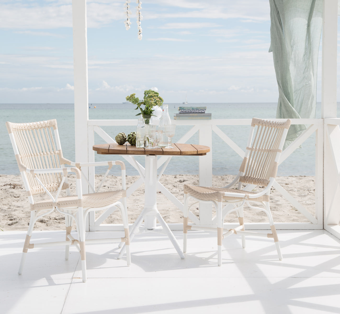 Dette er et livsstilsbillede af Nicole Cafe Table i white aluminium frame white ArtFibre. Med Piano-stole på et dæk ved havet.