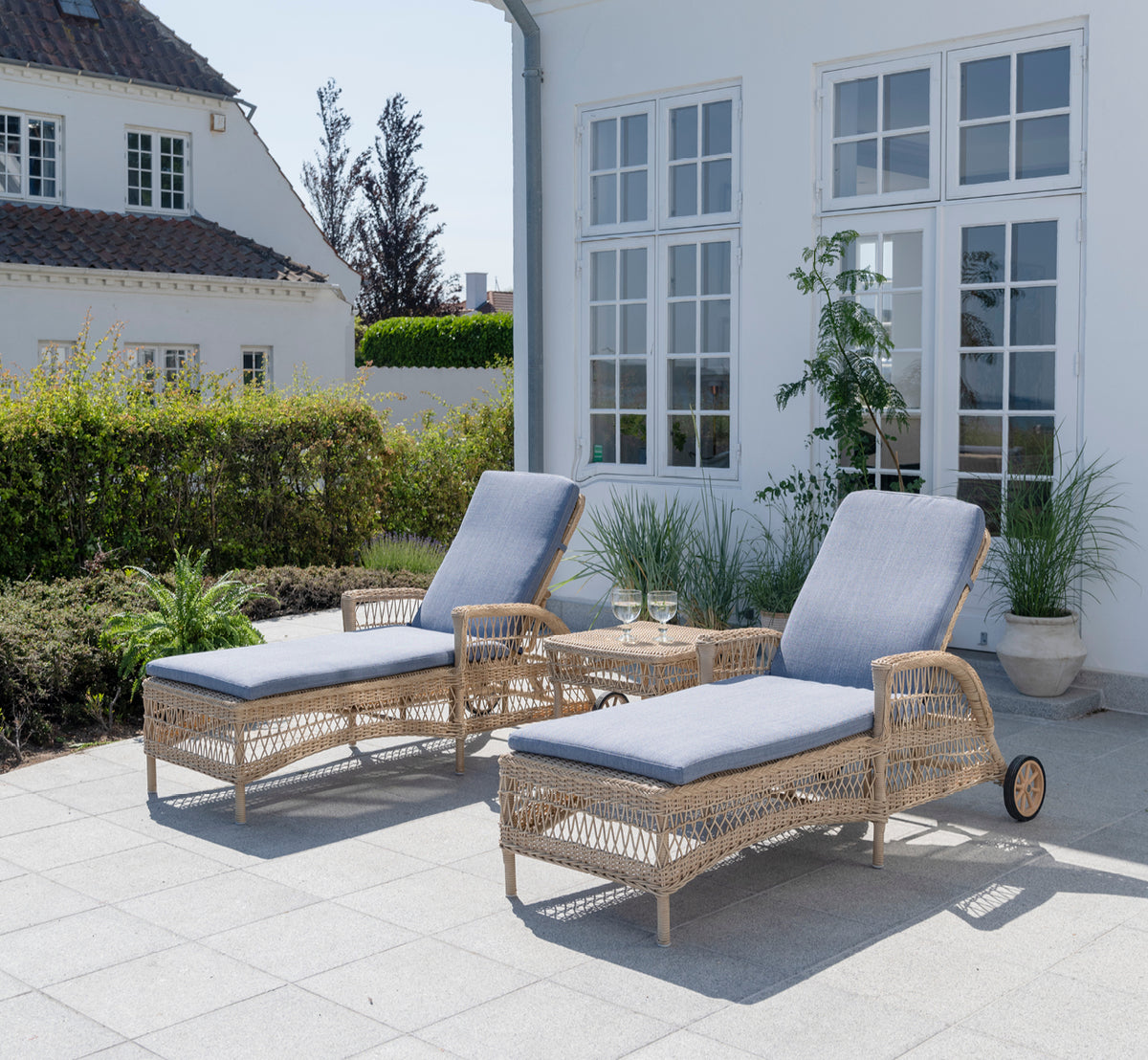 Dette er et livsstilsbillede af Daisy Solseng i natural ArtFibre Aluminium, på en haveterrasse omgivet af frodige træer og potteplanter.