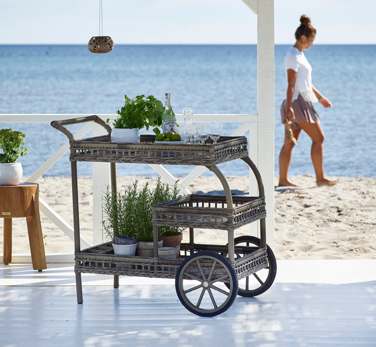Dette er et livsstilsbillede af James Serveringsvogn i antique grey artfibre aluminium, på en strandterrasse under en hvid pergola med havudsigt.