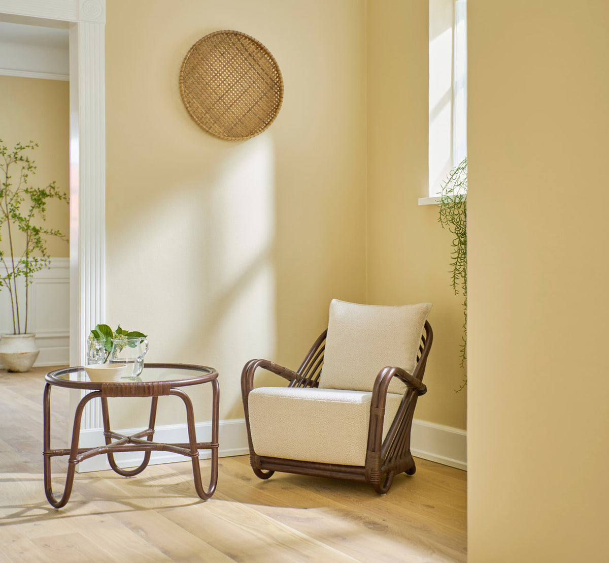 Dette er et livsstilsbillede af Charlottenborg Lænestol i brown rattan. Med sidebord i glas i et varmt gult hjørne.