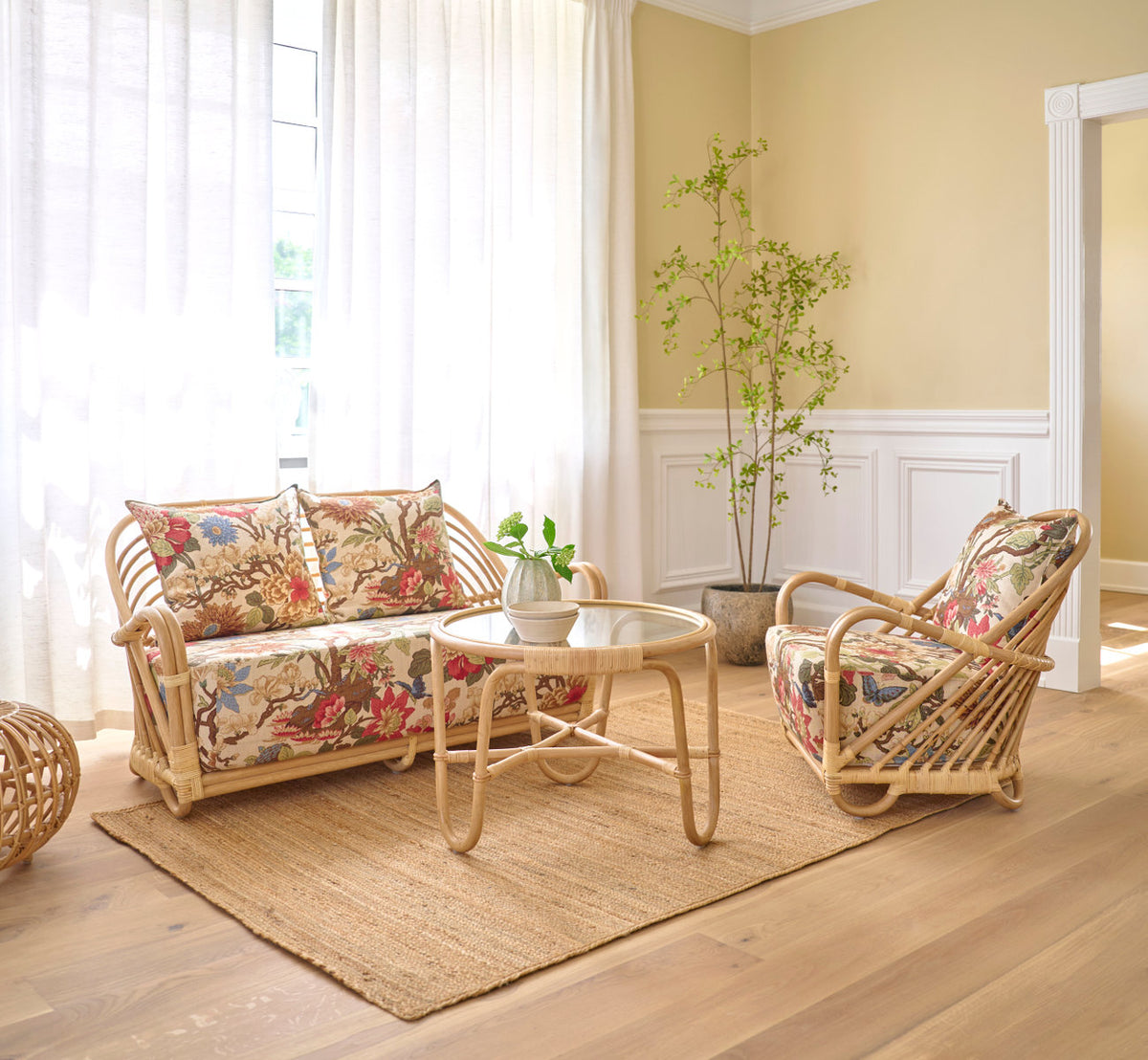 Dette er et livsstilsbillede af Charlottenborg Lænestol i polished natural rattan. Med blomstrede puder og sofabord på et jutetæppe.