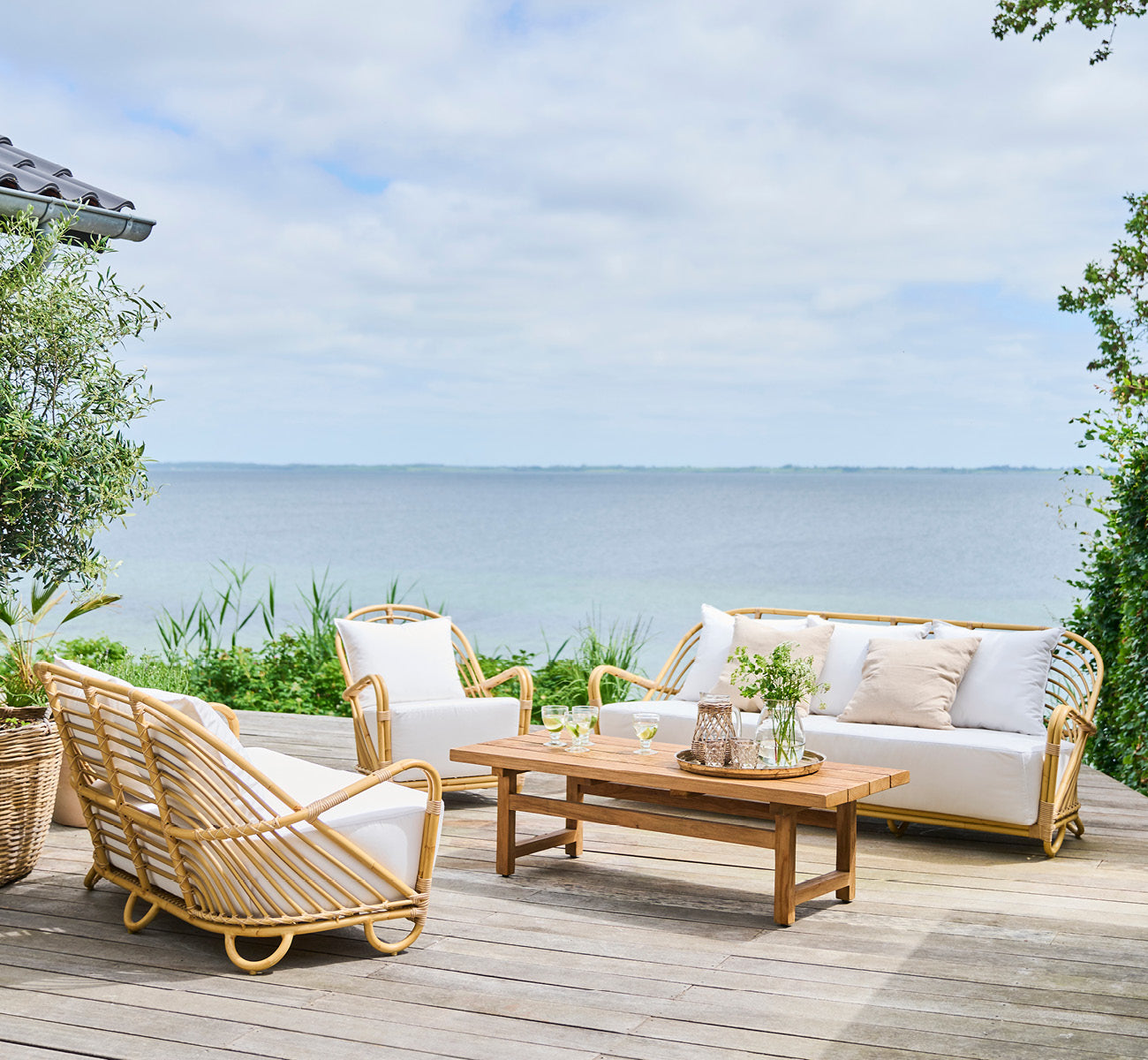 Dette er et livsstilsbillede af Charlottenborg Udendørs 2-personers sofa i natural Aluminium ArtFibre, på et udendørs trædæk med kystnær vegetation.