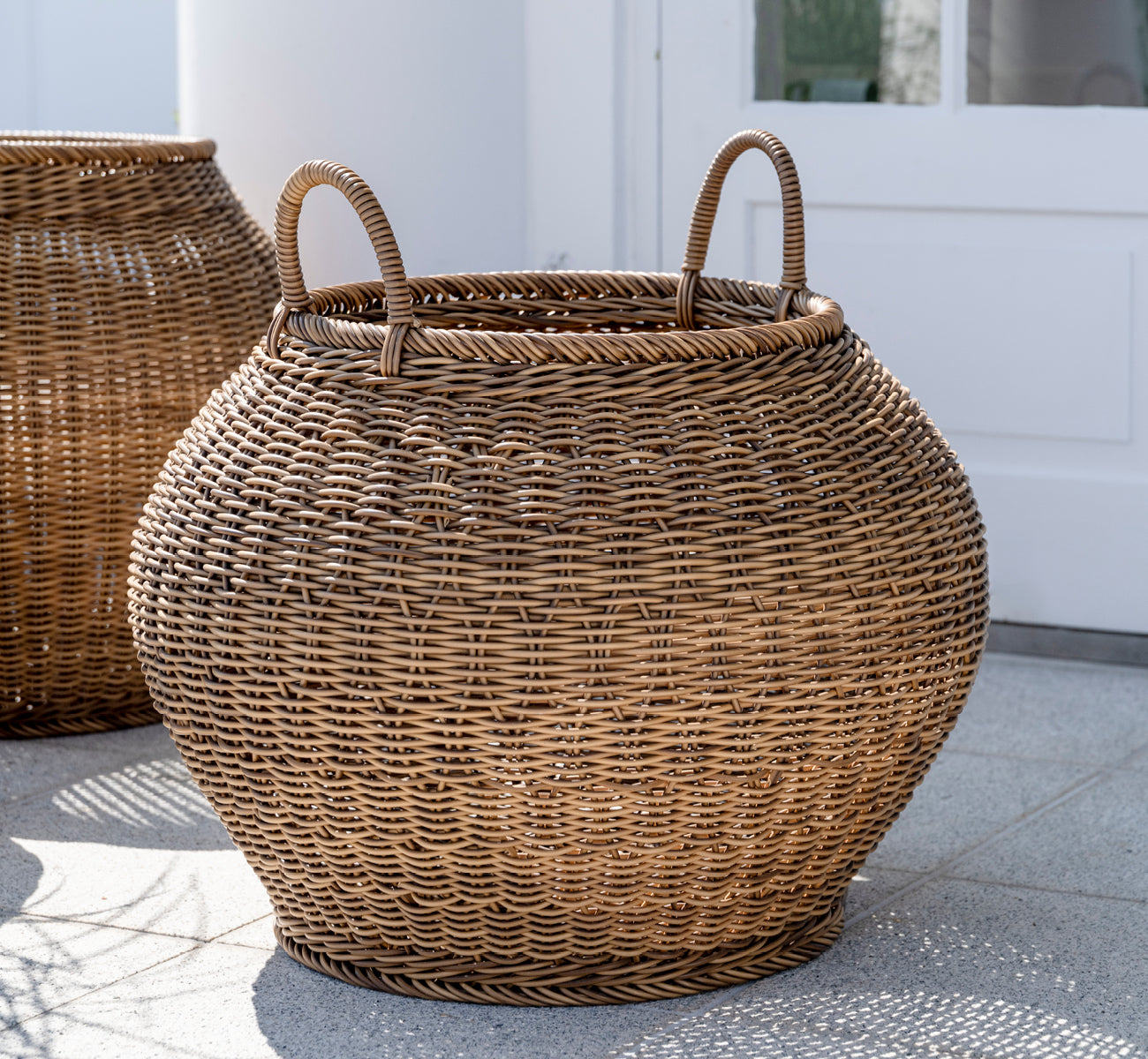 Dette er et livsstilsbillede af Smilla Kurv i chestnut core weave ArtFibre Aluminium, udendørs på en solbeskint terrasse med hvid mur og flisegulv.