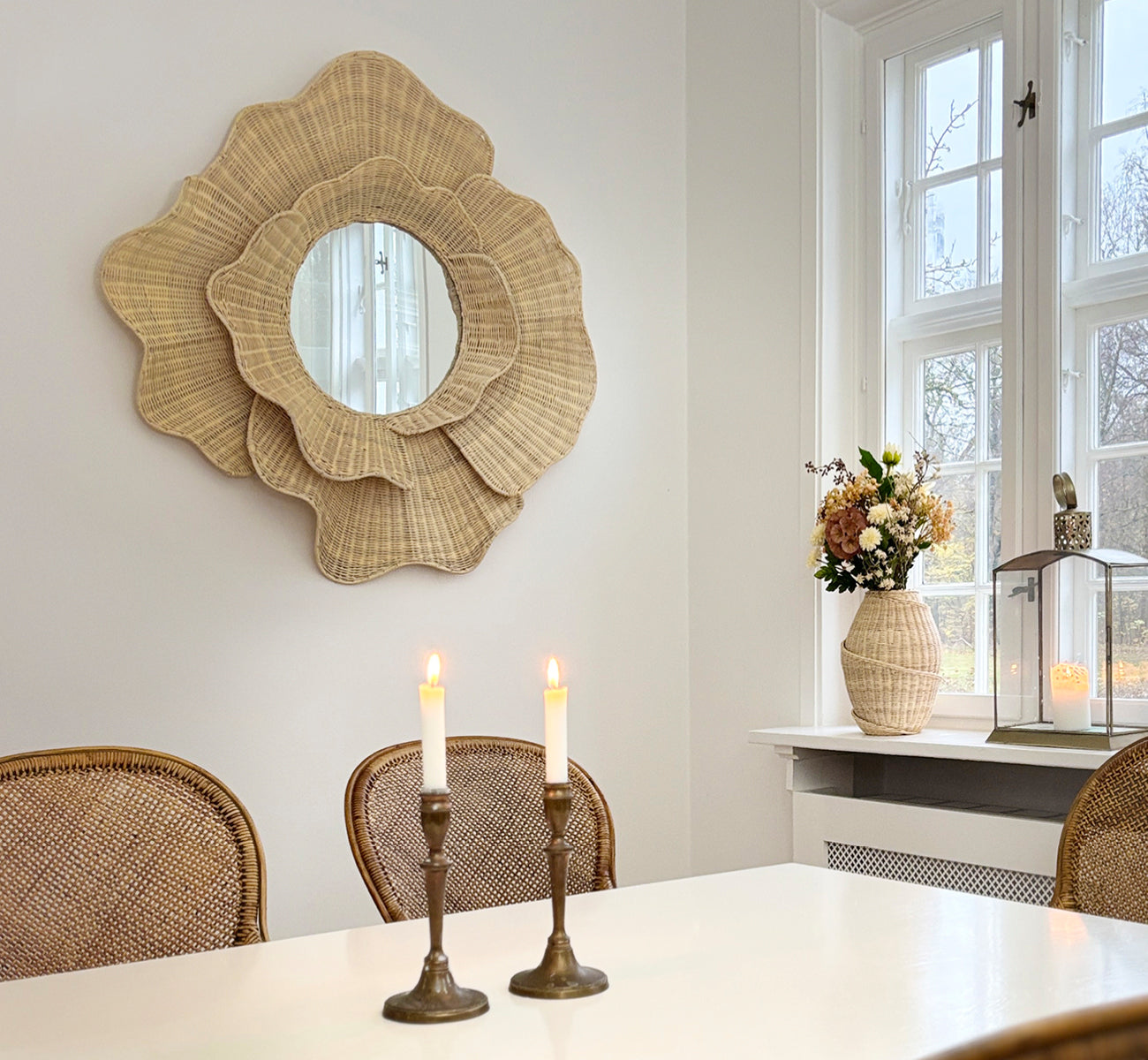 Dette er et livsstilsbillede af Floral Spejl i natural core weave iron frame, hængende i en lys spisestue med lysestager og naturligt vindueslys.