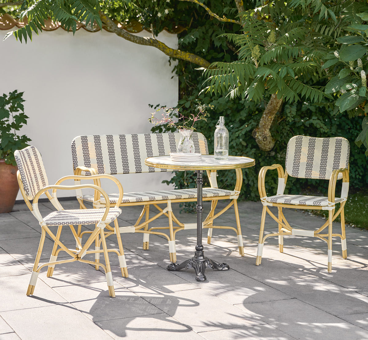 Dette er et livsstilsbillede af Bistro Exterior Stol i natural Aluminium ArtFibre, i et udendørs havecafé med et bistrobord og omkringstående træer.
